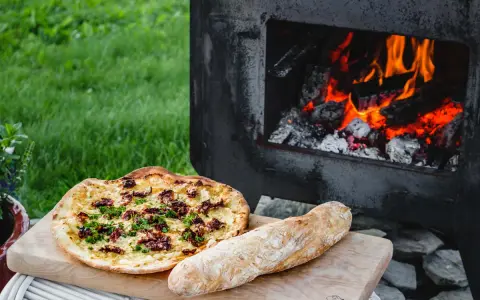 En pizza och ett bröd ligger på en skärbräda bredvid en pizzaugn med flammande ved.