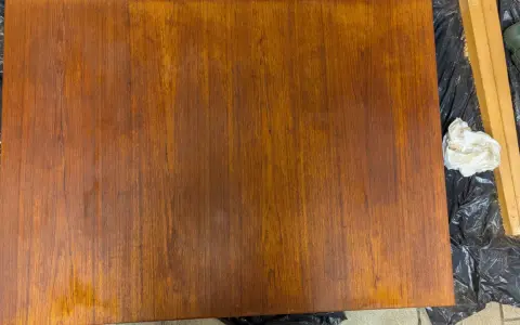Förbereder för att slipa teakbord. En bit trä ligger på svart plast, bredvid en slipmaskin och en pappersbit.