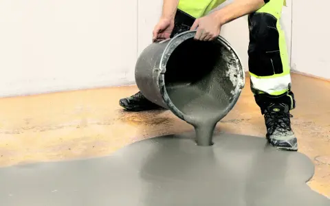 Person i varselkläder häller flytspackel golv ur en hink. Det gråa spacklet sprider sig över det gamla golvet.
