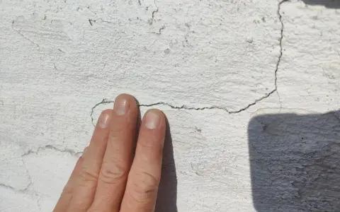 En hand pekar på sprickor i en vit, texturerad fasad.