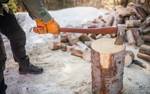 Person med yxa klyva ved i snöig skog. En hög med ved i bakgrunden.
