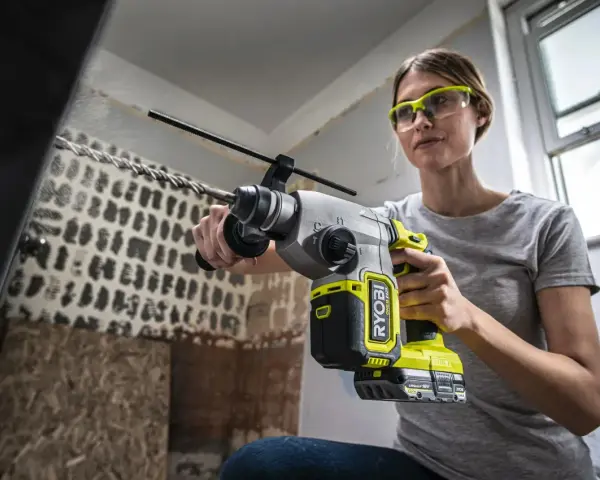 Kvinna med skyddsglasögon borrar i betongtak med en kraftfull slagborrmaskin. Damm virvlar upp från arbetet.