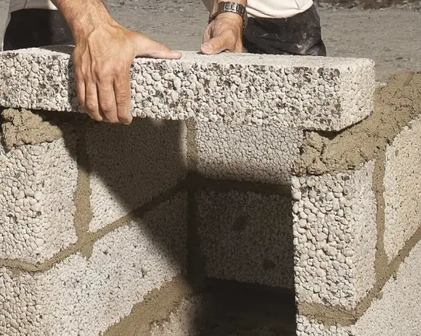 Händer lägger ett mura lecablock på en grund av cement. Byggnationen pågår utomhus.