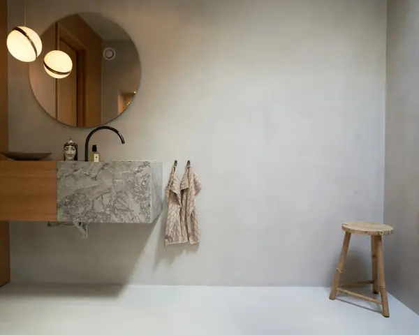 Modernt badrum med handfat i marmor och väggar av microcement. Enkel träpall och rund spegel.