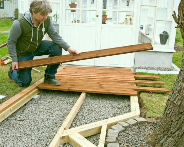 Man bygger trall vid ett växthus. Han håller en träbräda redo att läggas på plats.