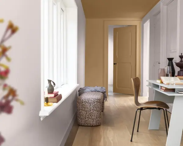 Ett rum med ljusa väggar och en dörr som är målad i en varm, gyllene ton. En vit fönsterbräda med böcker och en vas. En bänk med blommigt tyg och en trästol vid ett skrivbord.
