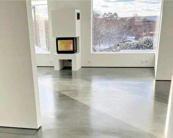 Ett modernt vardagsrum med en öppen spis och stora fönster som vetter mot en snötäckt trädgård. Golvet är ett vackert polerat betonggolv som reflekterar ljuset.