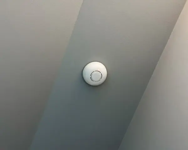 En vit, rund, uppkopplad brandvarnare sitter monterad i ett tak med två olika gråa nyanser.