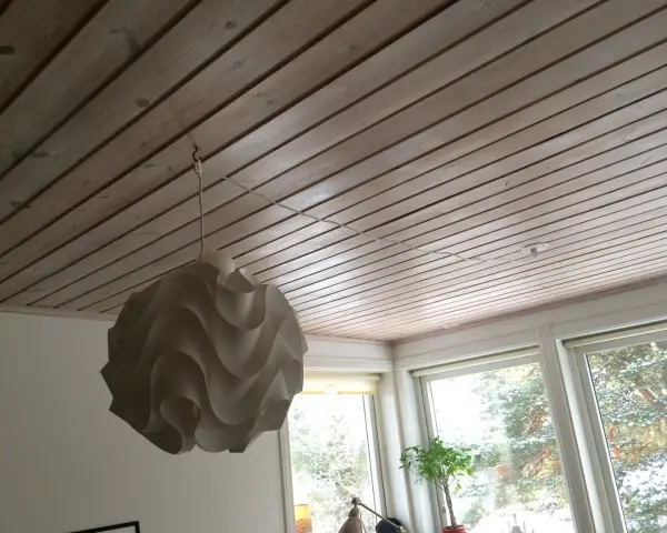 En vit taklampa hänger från en krok i ett träpanelat tak. En tunn sladd löper från lampan längs taket mot ett uttag.