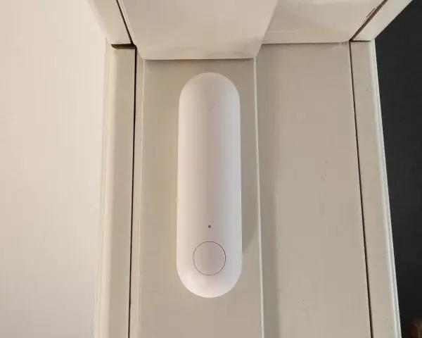 En vit, avlång sensor med en knapp sitter monterad på en ljusbeige dörrkarm. Den är en del av ett Aqara Thread-system för smart hem.