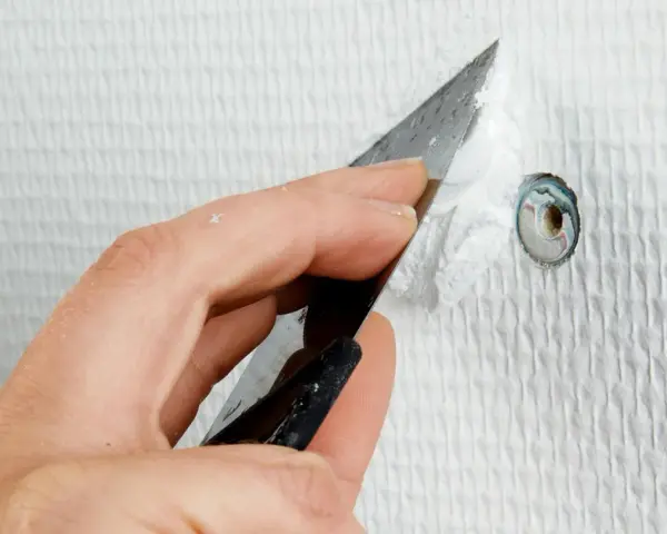 Hand spacklar ett litet hål i en vit, strukturerad vägg med en spackelspade.