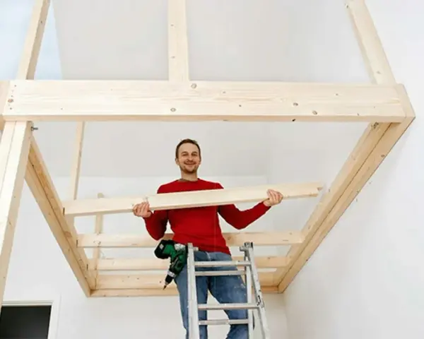 Man på stege bygger ett loft av trä. Han håller en träbit och ler.