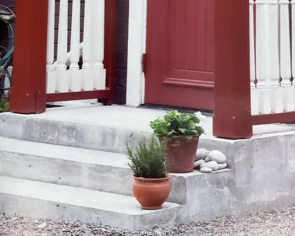 Nygjuten betongtrappa med röda stolpar och vita baluster. Krukor med växter och stenar pryder trappsteget.