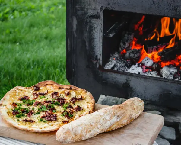 En pizza och ett bröd ligger på en skärbräda bredvid en pizzaugn med flammande ved.