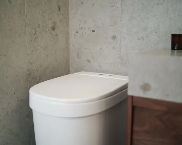 Modernt badrum med en vit toalett utan avlopp, grå kaklade väggar och ett handfat i betong.