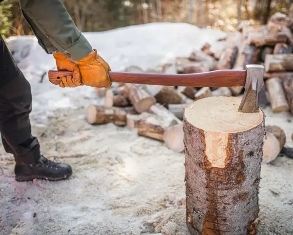 Person med yxa klyva ved i snöig skog. En hög med ved i bakgrunden.