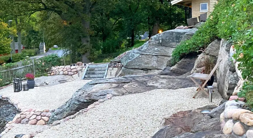 En stenlagd uteplats med grusgångar, stenmurar och trappor som leder upp mot ett hus. En inspiration för en naturlig och robust utomhusmiljö.