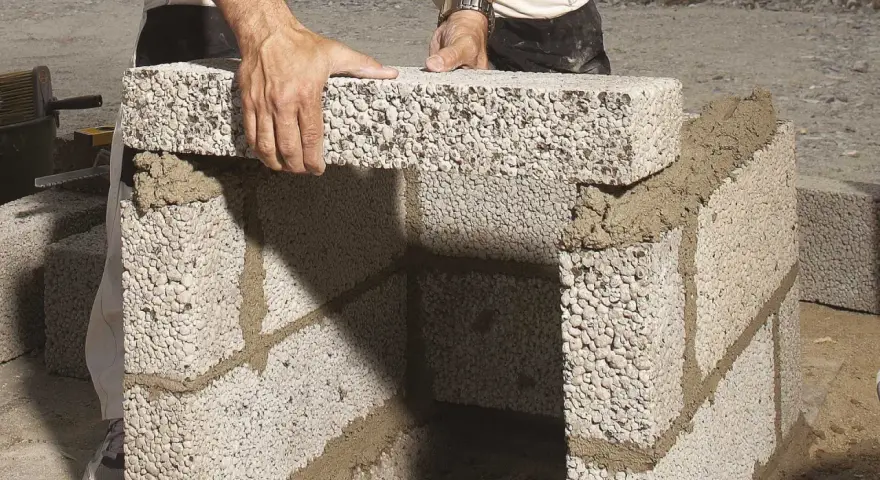 Händer lägger ett mura lecablock på en grund av cement. Byggnationen pågår utomhus.