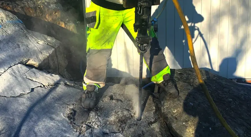 Arbetare med reflexkläder borrar i berg med en kraftig borrhammare. Dammet yr runt borren.