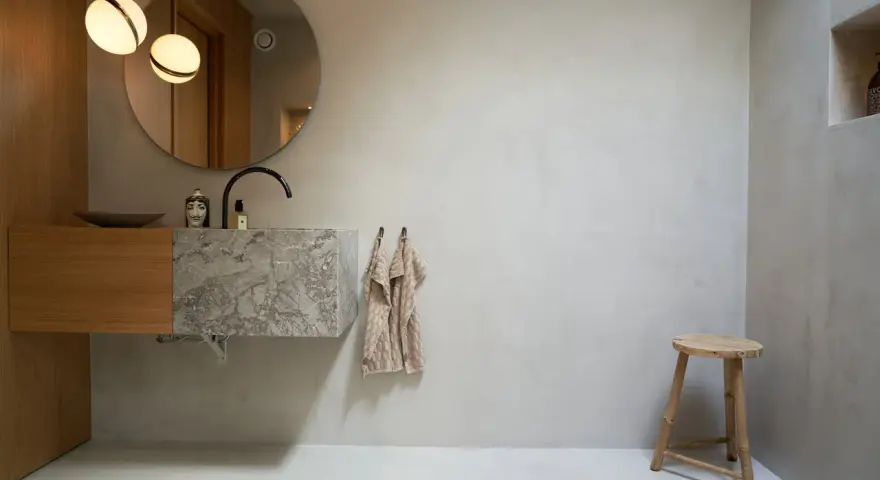 Modernt badrum med handfat i marmor och väggar av microcement. Enkel träpall och rund spegel.