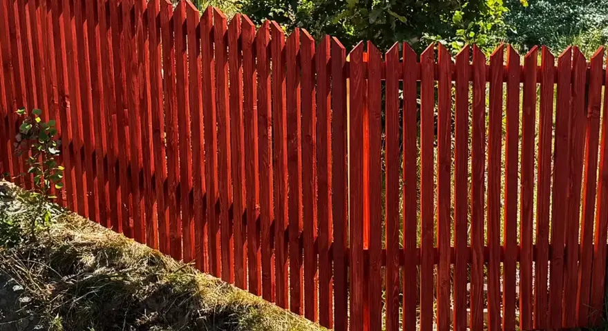 Rött trästaket med spetsiga toppar ger fin staket inspiration. Grönskande träd i bakgrunden.