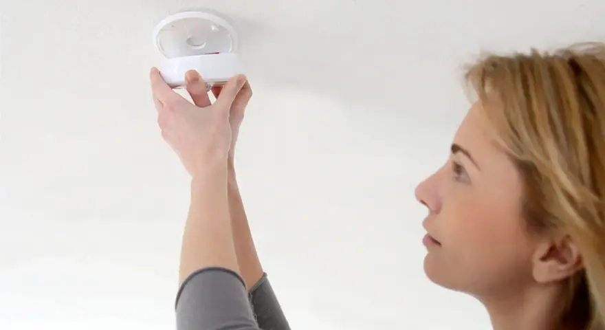 En kvinna installerar en brandvarnare. Dessa brandvarnare som pratar med varandra är viktiga för säkerheten.