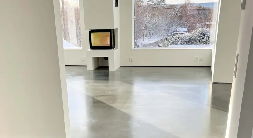 Ett modernt vardagsrum med en öppen spis och stora fönster som vetter mot en snötäckt trädgård. Golvet är ett vackert polerat betonggolv som reflekterar ljuset.