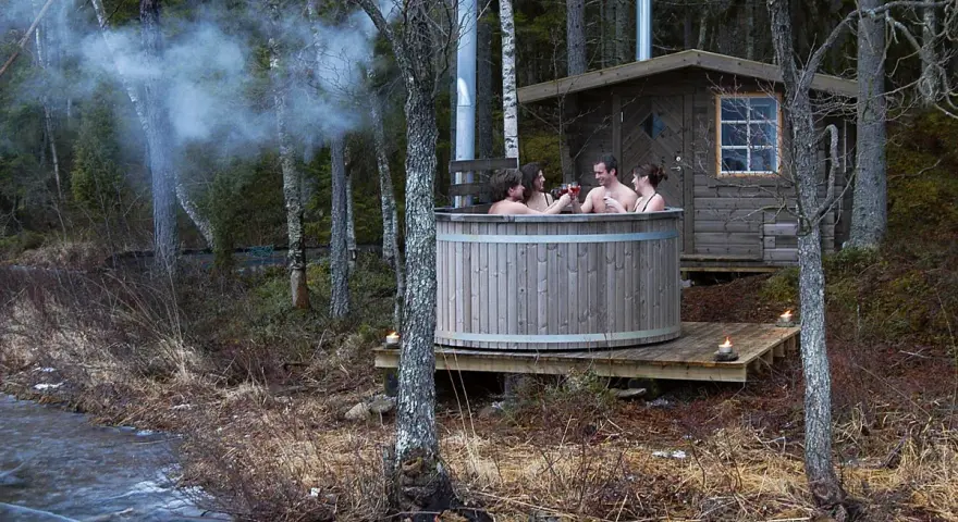 Fyra vänner njuter av en badtunna vid en sjö. Röken stiger från skorstenen, en mysig stund att bygga badtunna för.