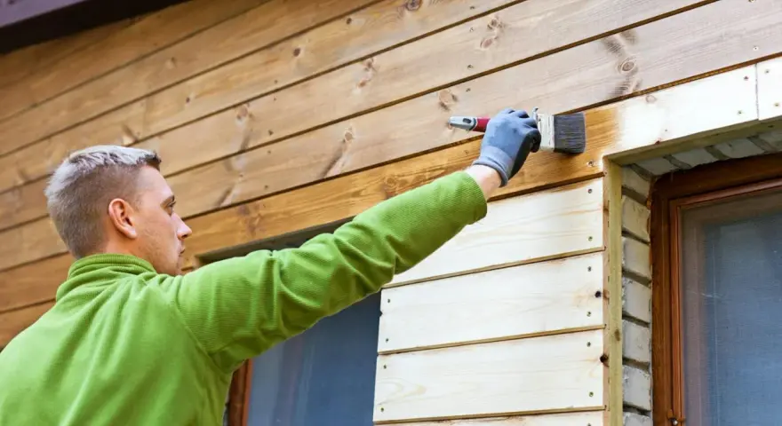 Man målar tryckimpregnerat trä med en pensel. Han har en grön tröja och grå handskar.