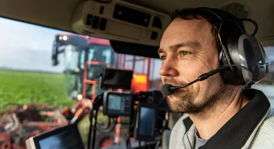 En lantbrukare i förarhytten på en skördetröska, med hörlurar som liknar Peltor WS Alert XP vs XPI, fokuserar på arbetet på fältet.