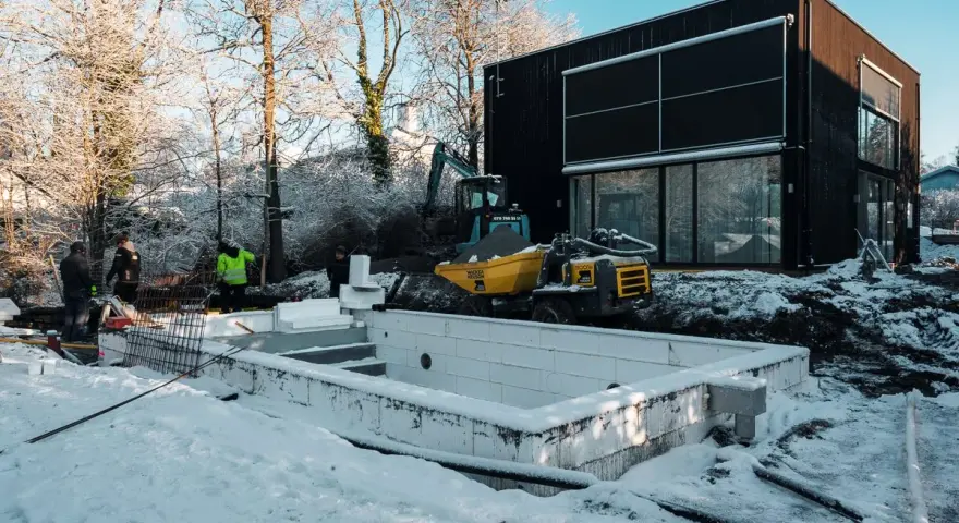 Snö täcker byggarbetsplatsen där en utbyggnad av husets långsida pågår. En grävmaskin och en dumper syns vid en nybyggd poolgrund.
