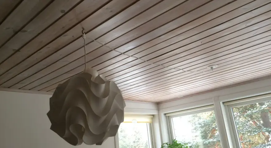 En vit taklampa hänger från en krok i ett träpanelat tak. En tunn sladd löper från lampan längs taket mot ett uttag.