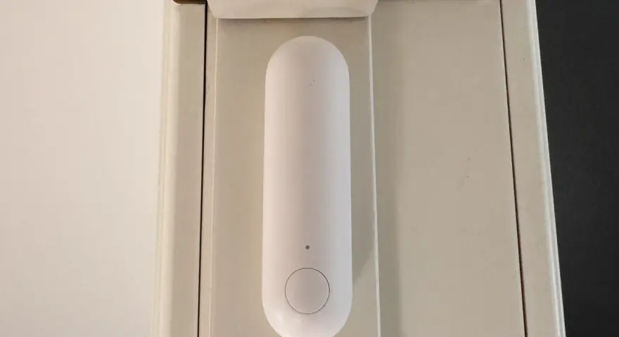 En vit, avlång sensor med en knapp sitter monterad på en ljusbeige dörrkarm. Den är en del av ett Aqara Thread-system för smart hem.