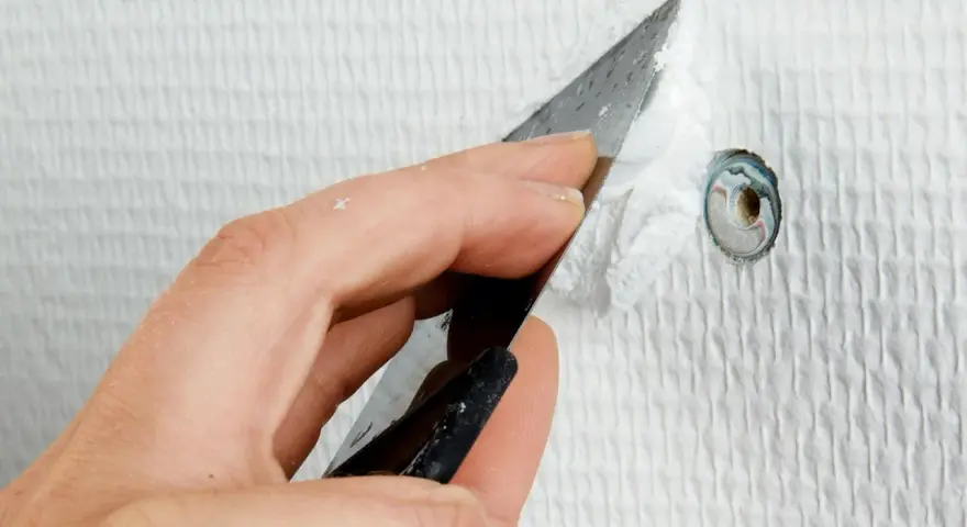 Hand spacklar ett litet hål i en vit, strukturerad vägg med en spackelspade.