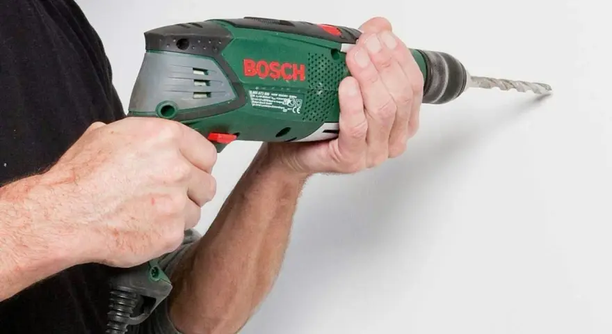 Händerna håller en grön Bosch-borrmaskin som borrar i en vit vägg.
