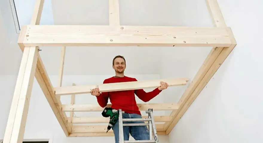 Man på stege bygger ett loft av trä. Han håller en träbit och ler.