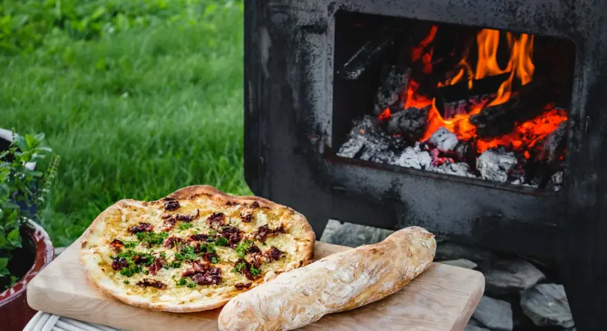 En pizza och ett bröd ligger på en skärbräda bredvid en pizzaugn med flammande ved.