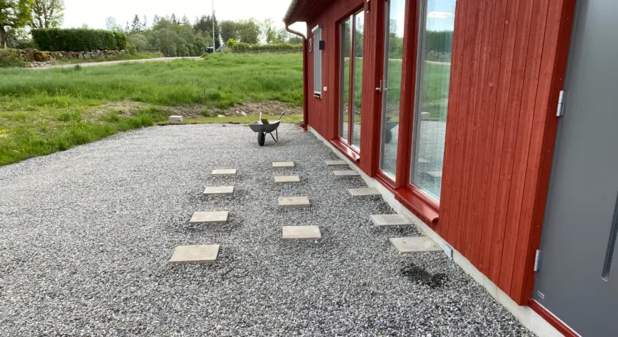 Stigande steg av altan plintar eller plattor leder genom grusgången fram till ett rött hus med stora fönster.
