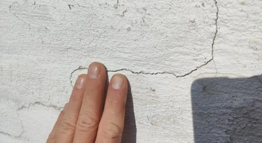 En hand pekar på sprickor i en vit, texturerad fasad.