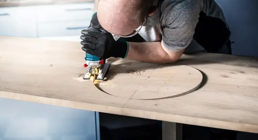 Man med skyddsglasögon och handskar använder sticksåg för att såga rakt i en träskiva.