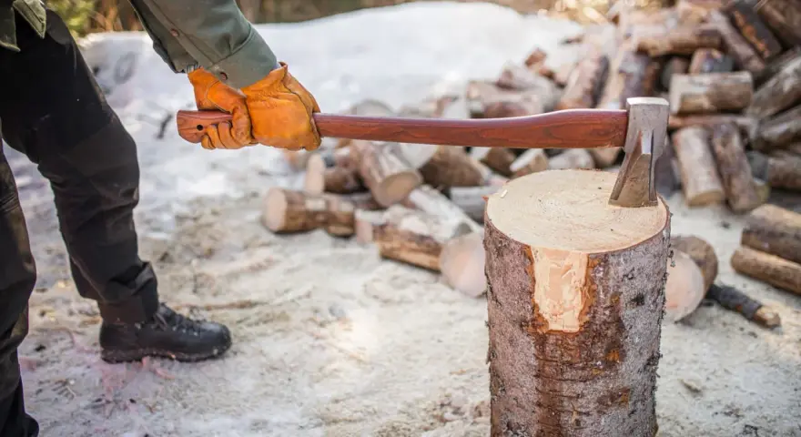 Person med yxa klyva ved i snöig skog. En hög med ved i bakgrunden.