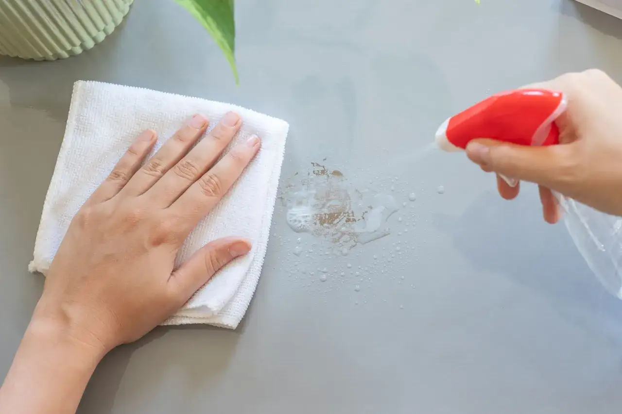 En hand med en vit trasa torkar bort envisa tejprester från en grå yta. En sprayflaska sprutar rengöringsvätska.