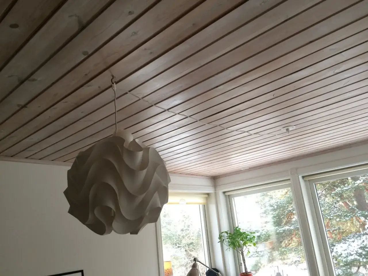 En vit lampskärm hänger från ett tak av träpanel. En tunn sladd löper från lampskärmen och fästs i taket.
