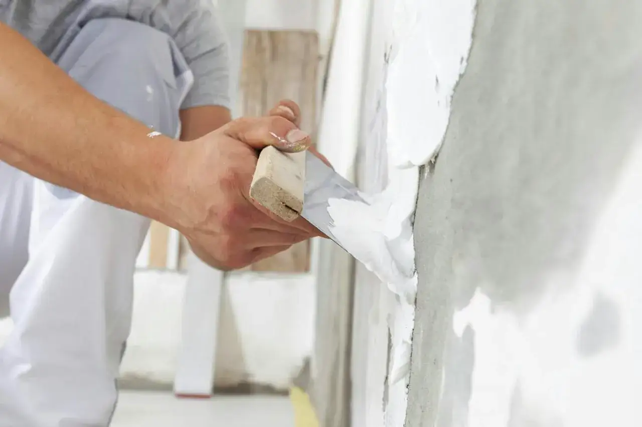 Händer med spackel och spackelspade applicerar putsbruk inomhus på en vägg.