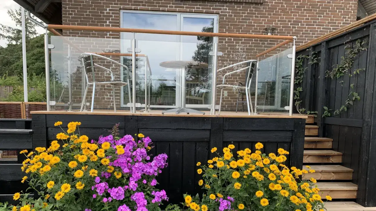Vacker altan med glasräcke och trämöbler, omgiven av blommor. Ett modernt altan staket inspiration för din uteplats.