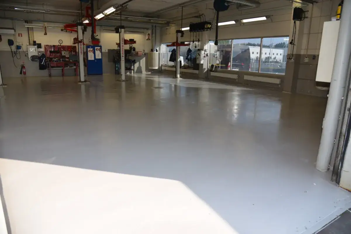 Ett ljust, nymålat garagegolv som reflekterar ljuset. Verktyg och billyftar syns i bakgrunden, redo för arbete.