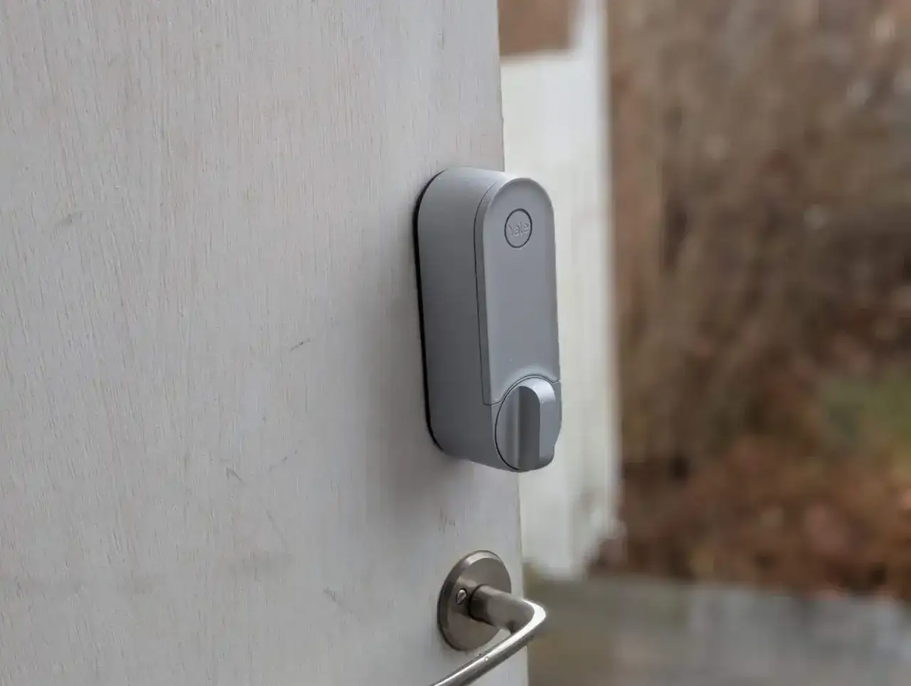 Ett modernt Yale nfc l&aring;s monterat p&aring; en vit d&ouml;rr. D&ouml;rren &auml;r p&aring; gl&auml;nt och visar en suddig bakgrund utomhus.