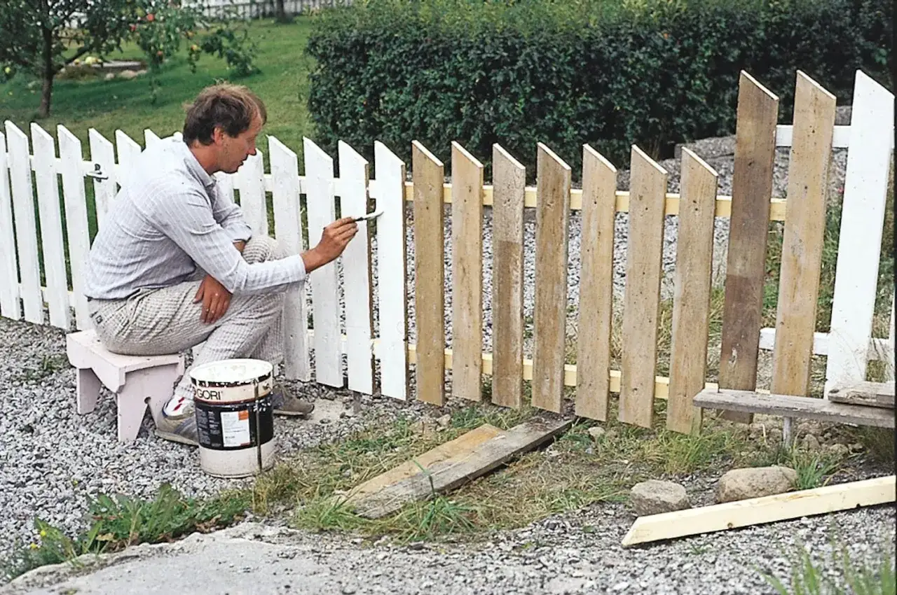 Man målar ett staket utomhus med vit färg. En burk med olja ek står bredvid.