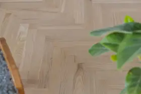 Ett nyslipat parkettgolv i fiskbensmönster, redo att skyddas. En grön växt och en del av en stol syns i bild.