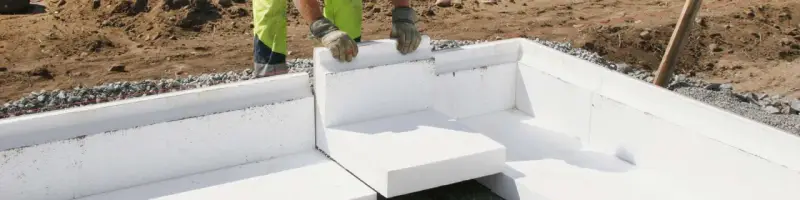 Arbetare monterar isolerblock för att gjuta platta garage. Grus och jord syns runt grunden.